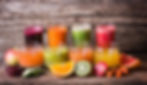 Assorted fruit and vegetable juices in glasses on a wooden table, surrounded by fresh produce like oranges, kiwis, carrots, and apple.
